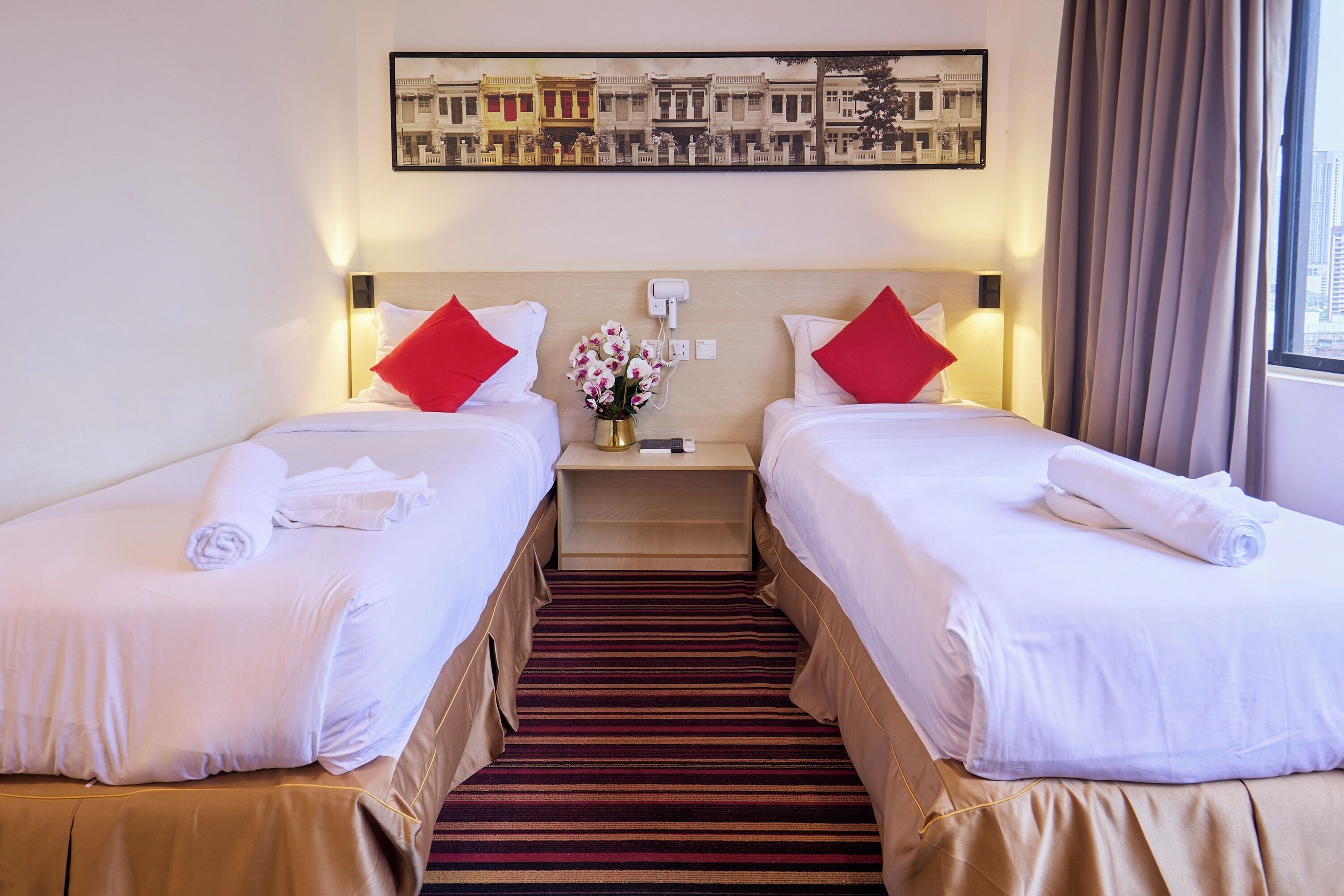 A hotel room with two beds, each with white linens and red pillows, a wooden headboard, a nightstand with a vase of flowers, and a cityscape painting above the headboard.