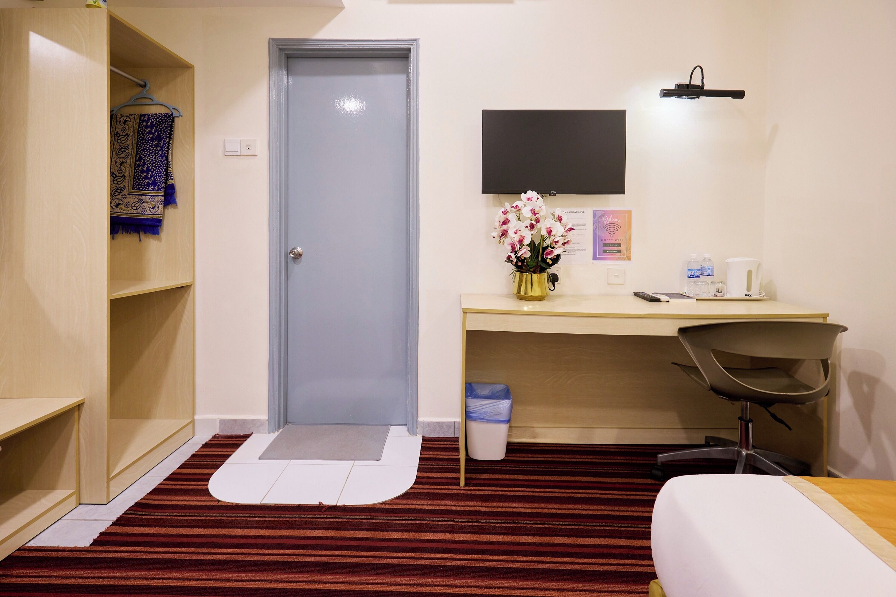Une chambre d’hôtel équipée d’un bureau, d’une chaise et d’un lit. On y trouve une porte, une petite poubelle et un téléviseur fixé au mur.
