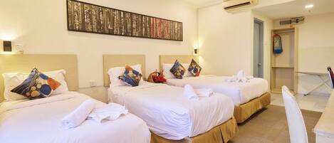 Habitación de hotel con tres camas, cada una con ropa de cama blanca y almohadas de colores. En la pared, un cuadro abstracto grande, un equipo de aire acondicionado y un escritorio pequeño con una silla.