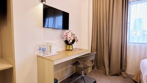 Een hotelkamer met een houten bureau, een stoel, een aan de muur bevestigde televisie, een vaas met bloemen en een raam met gordijnen.