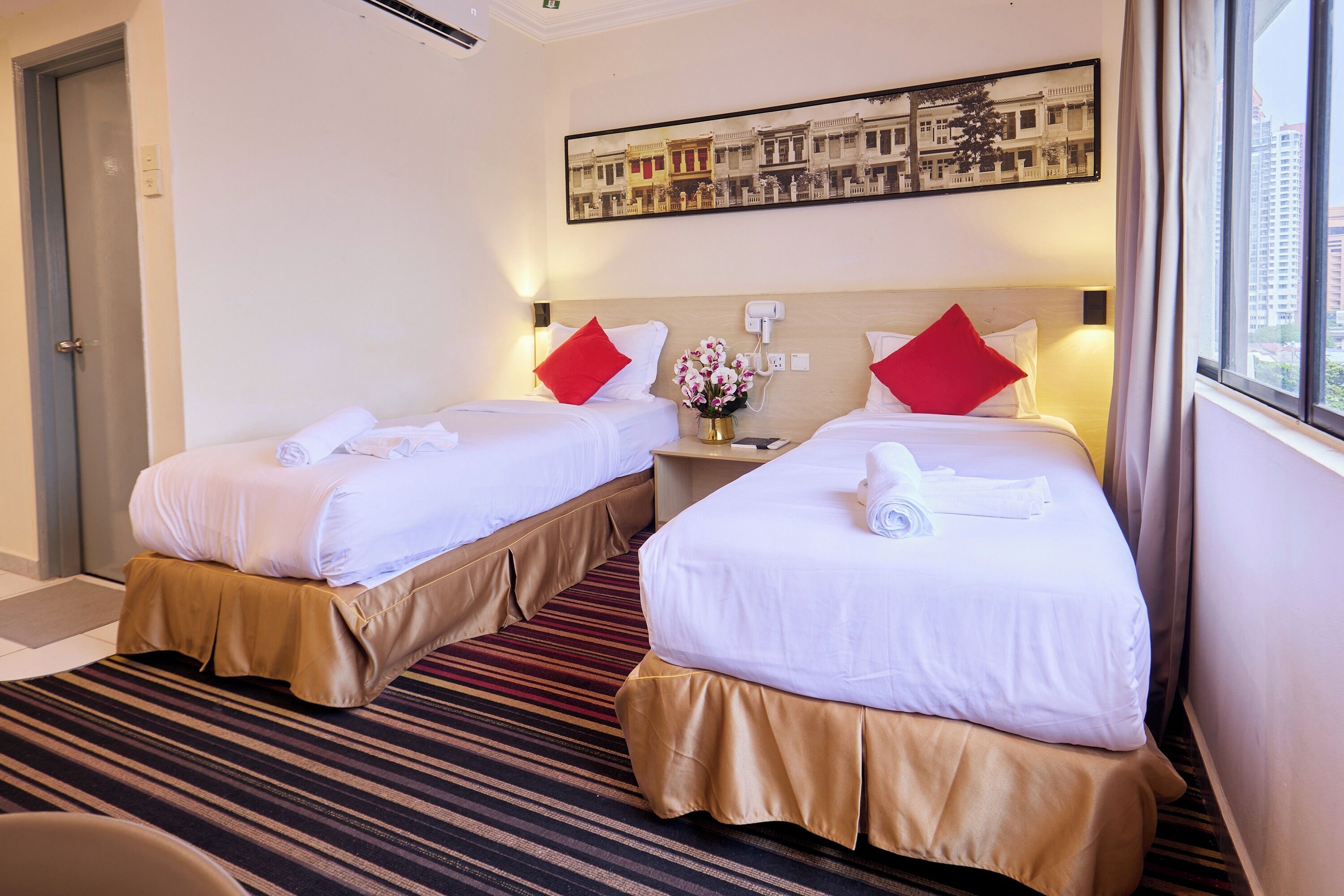 A hotel room with two beds, each with white linens and red pillows, a small table with a vase of flowers, and a framed picture on the wall.