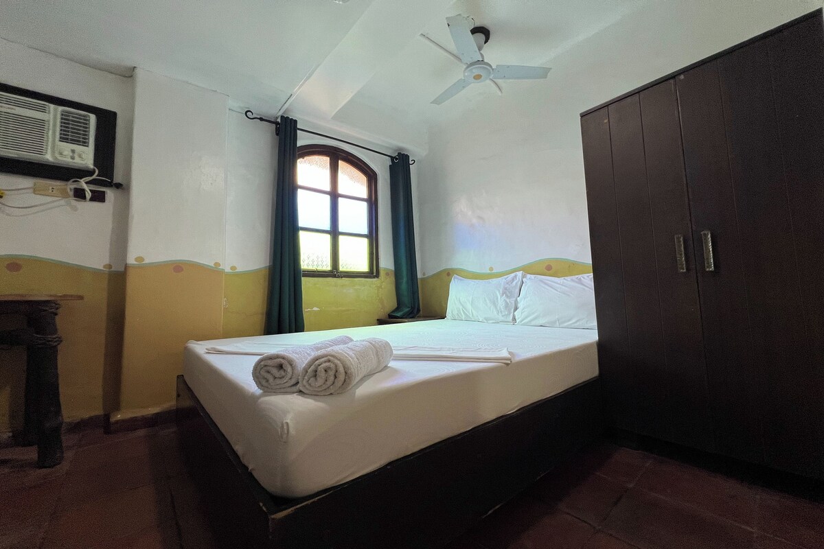 A single bed with a white mattress and pillows, a ceiling fan, a window with curtains, and a wardrobe.
