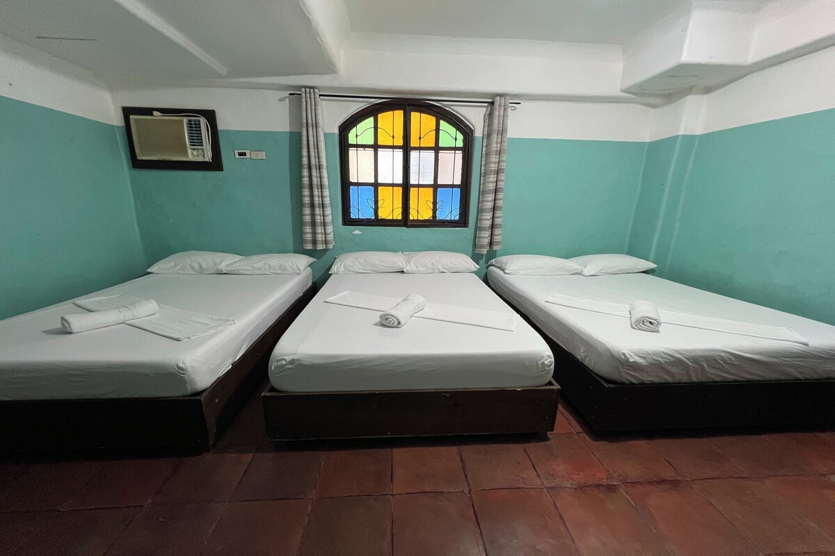 A room with three single beds, a window with a stained glass design, an air conditioning unit, and a wall-mounted light switch.