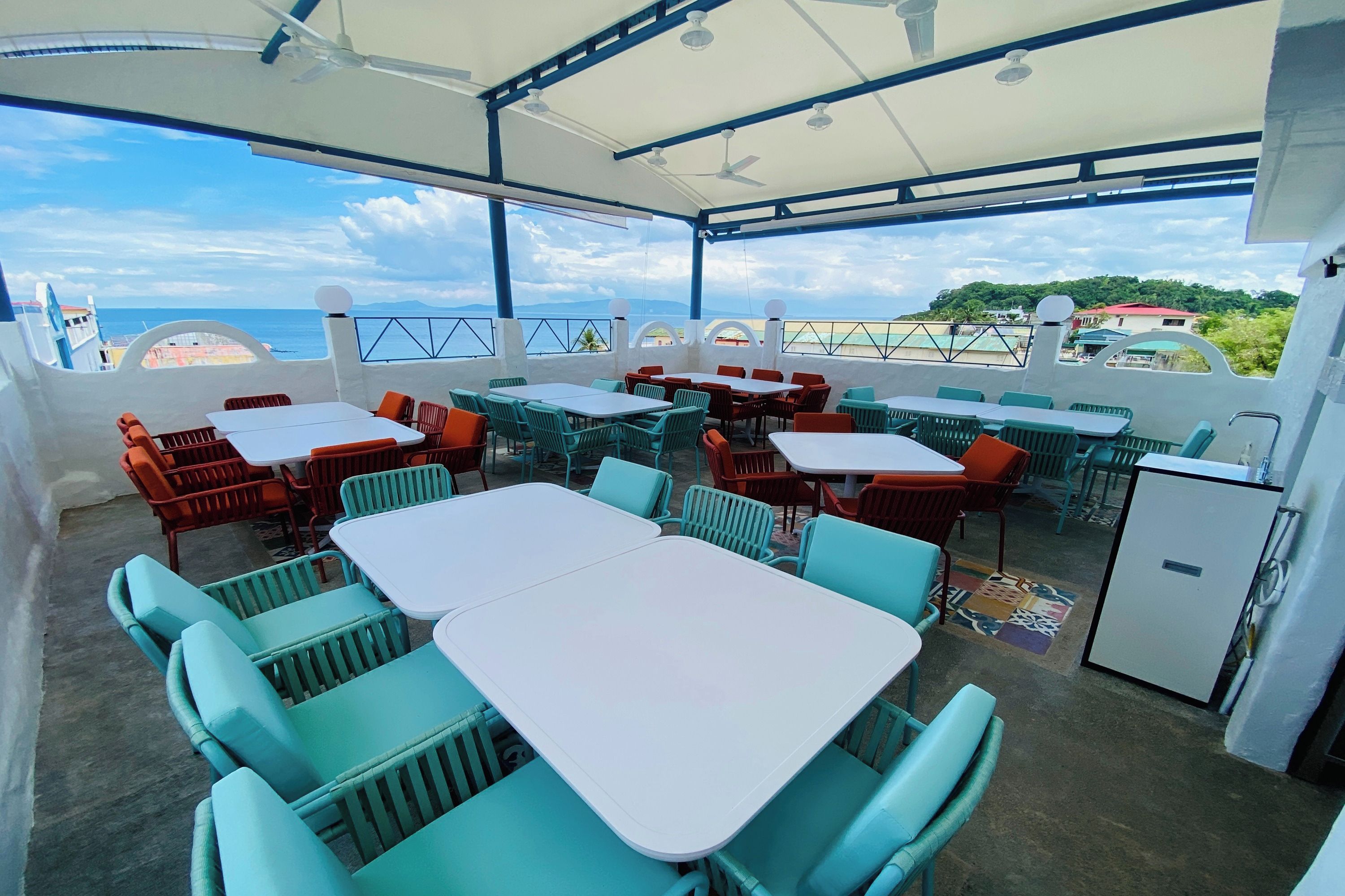 Un patio exterior con mesas y sillas, un ventilador de techo y vista al mar.