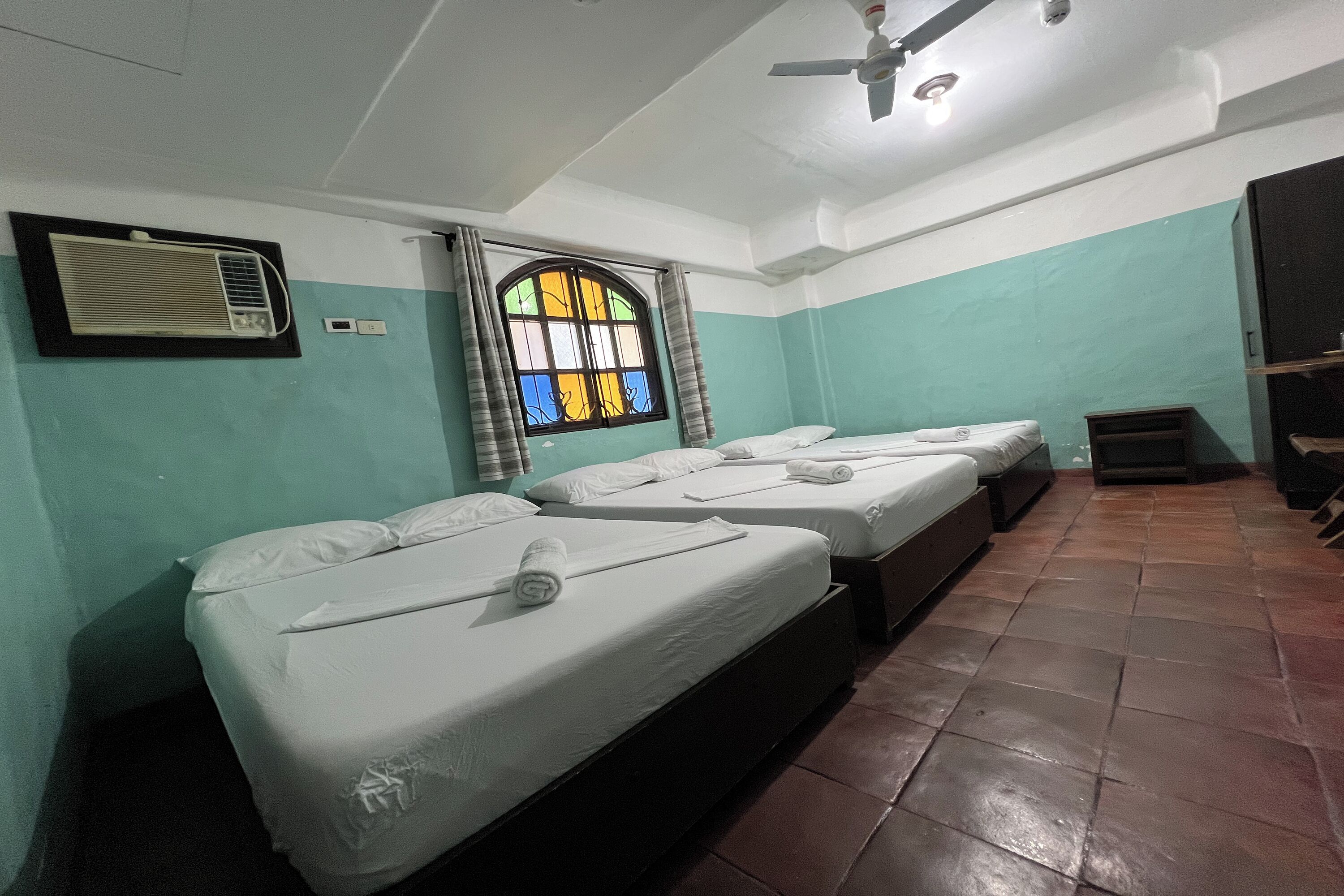 Una habitación con tres camas individuales, una ventana con cortinas, un aire acondicionado y un ventilador de techo.