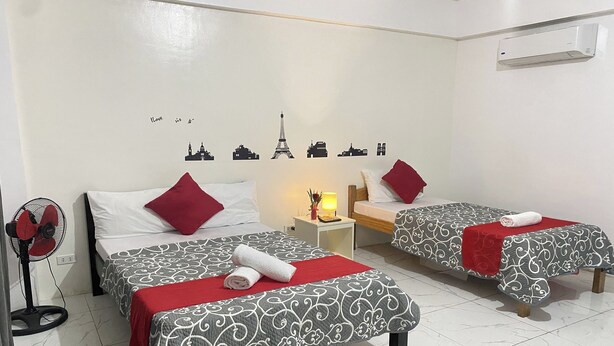 Una camera d'albergo con due letti, ognuno con una coperta a motivi decorativi e cuscini rossi. Si nota una decorazione murale con monumenti cittadini, una ventola da terra e un comodino con una lampada.