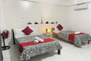 Habitación de hotel con dos camas, cada una con colcha estampada y almohadas rojas. Hay un aire acondicionado de pared y una mesita de noche con lámpara.