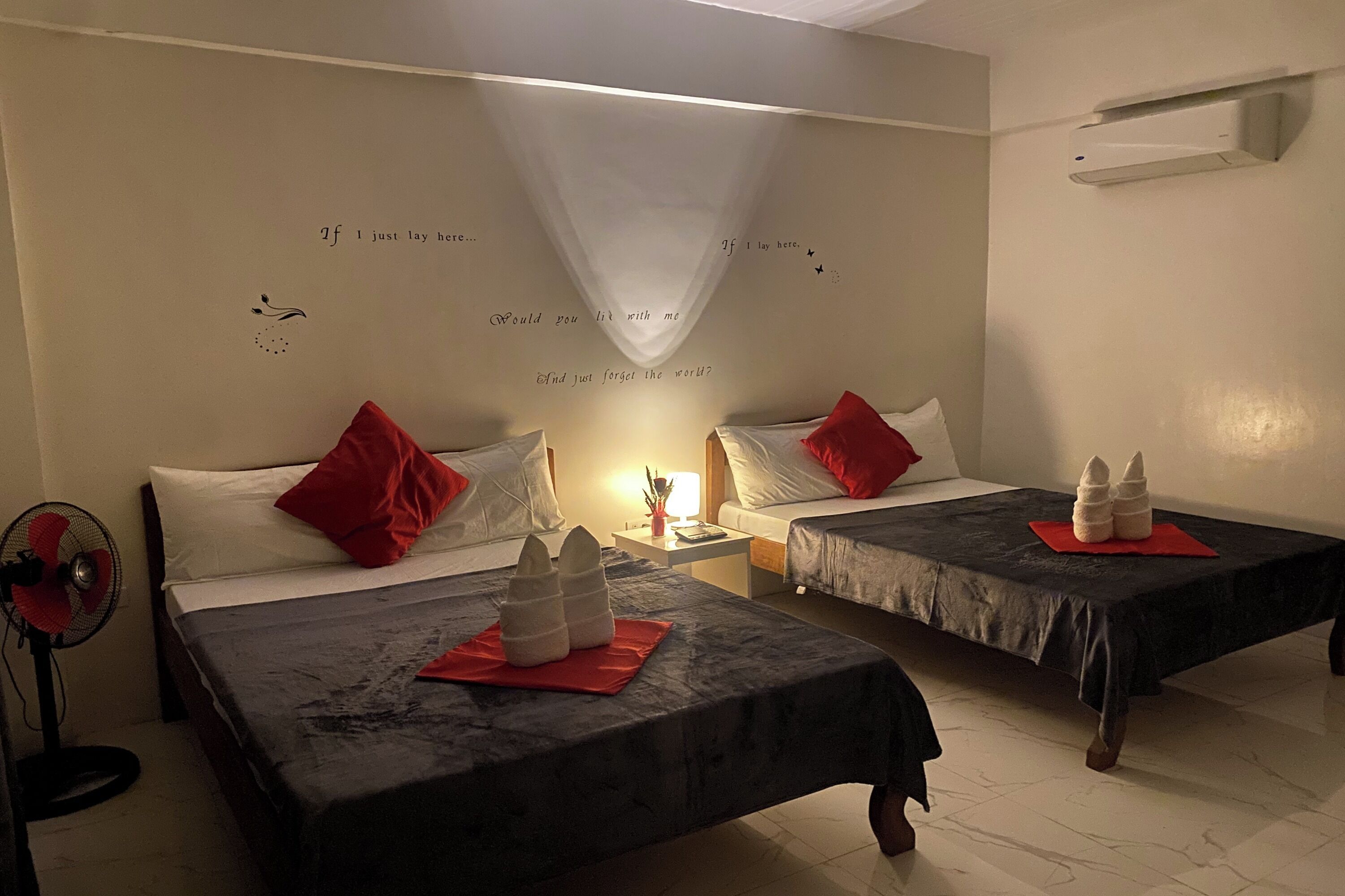 A hotel room with two beds, each with a red pillow and a white towel. There is a bedside table with a lamp and a small plant. The walls have decorative text and a white curtain.