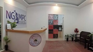 Un'area di accoglienza con un banco reception, una insegna a parete, una parete decorata, una zona salotto con una poltrona e un tavolino con una ventola.