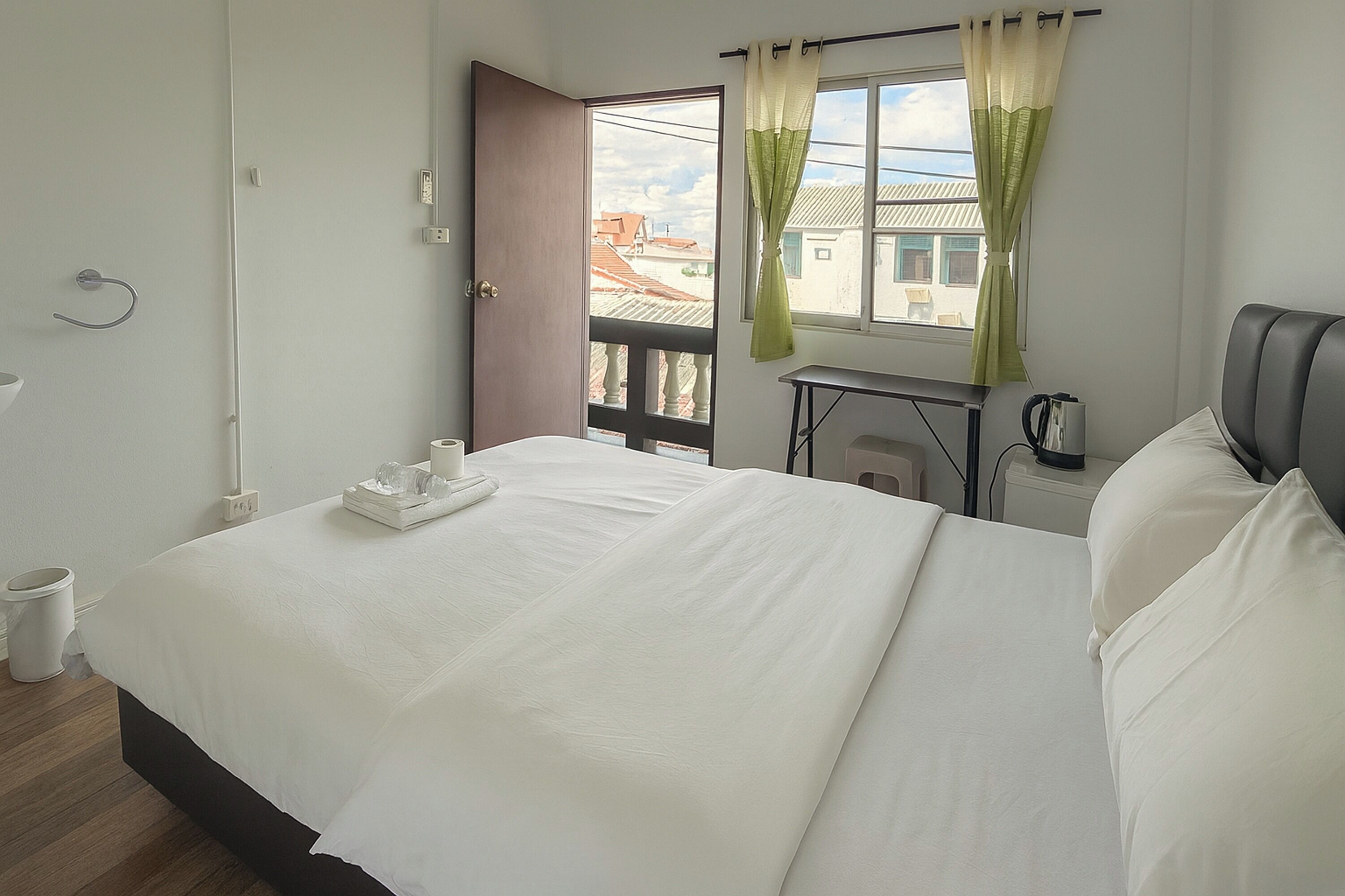 A neatly made bed with a tray of glasses and a cup, a small table with a kettle, and a view of buildings through an open door.
