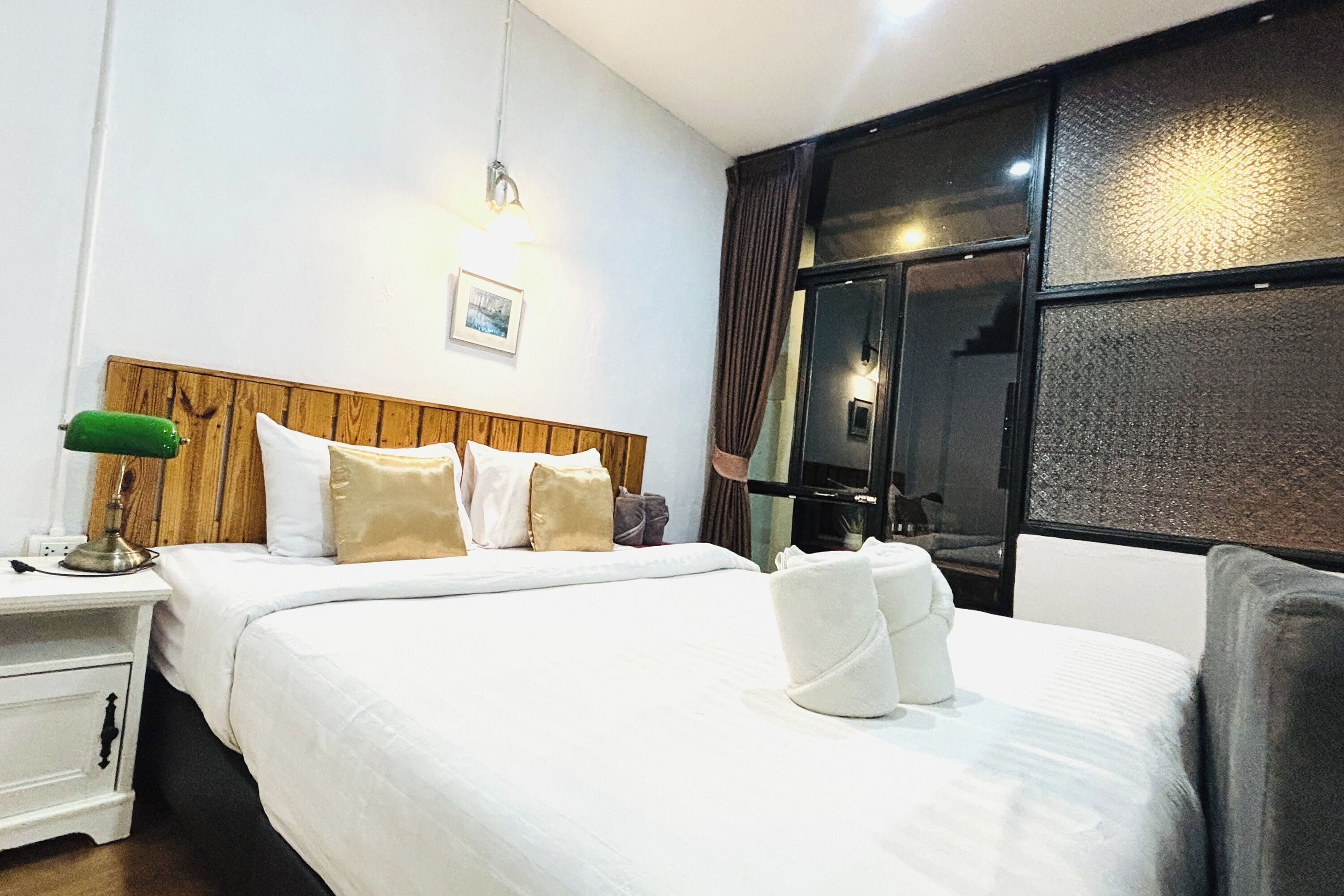 A neatly made bed with white linens, a wooden headboard, a bedside lamp, and a window with curtains.