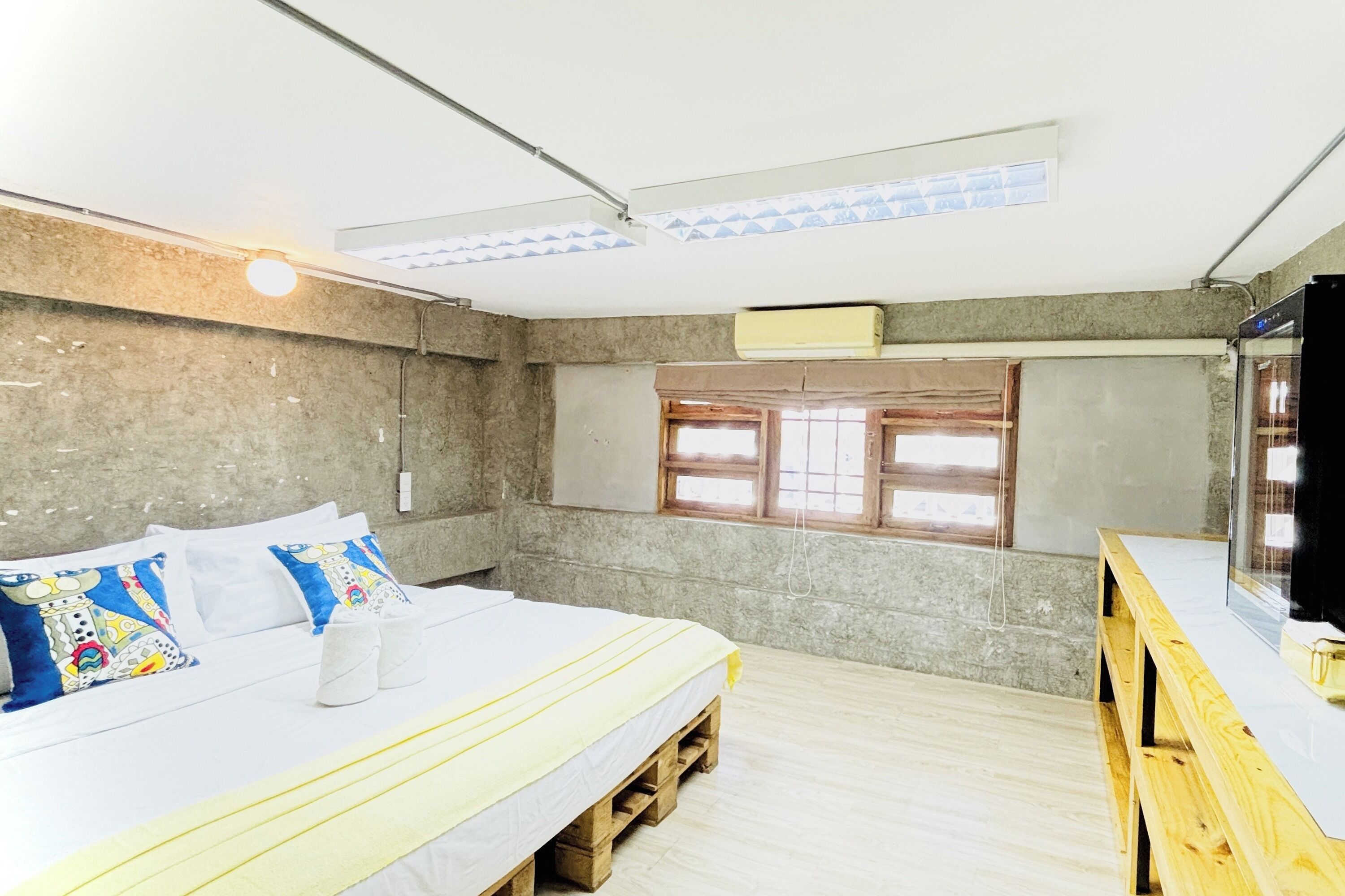A room with a bed, wooden floor, concrete walls, a window, and a TV.
