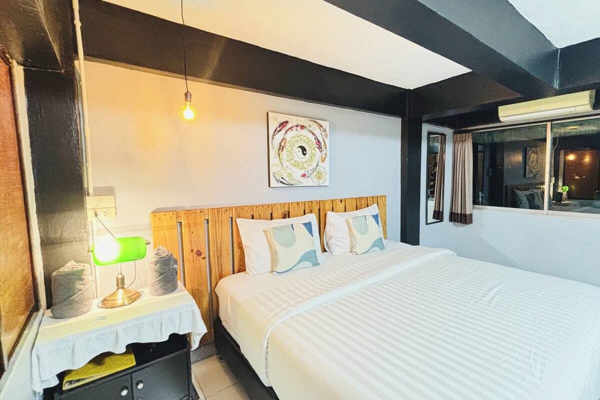 Habitación de hotel con cama, lámpara de noche, una pared con una obra de arte circular y una ventana con cortinas.