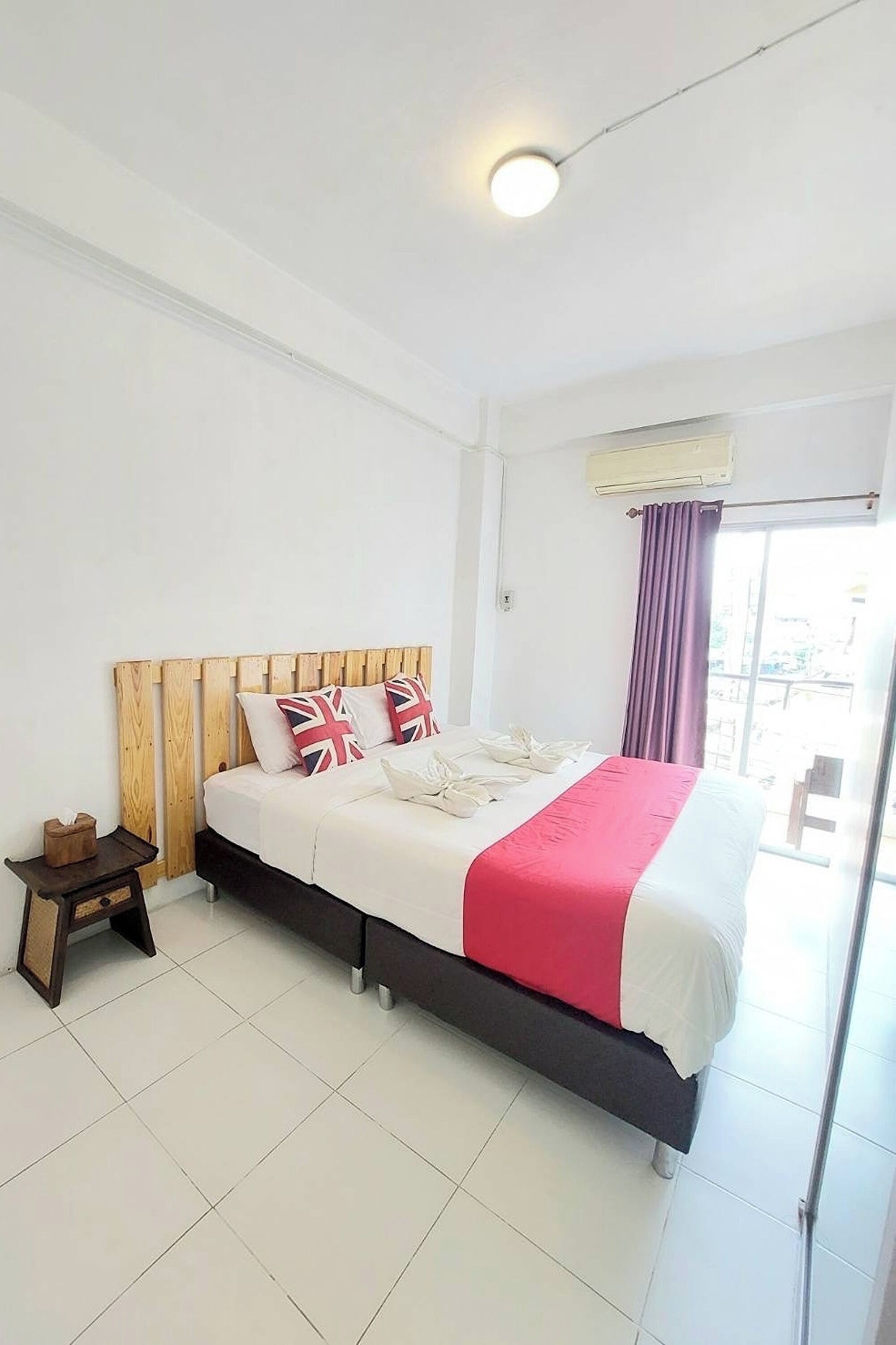 Una habitación de hotel con cabecera de madera, una cama con colcha roja y blanca, una mesita de noche con lámpara y una ventana con cortina.