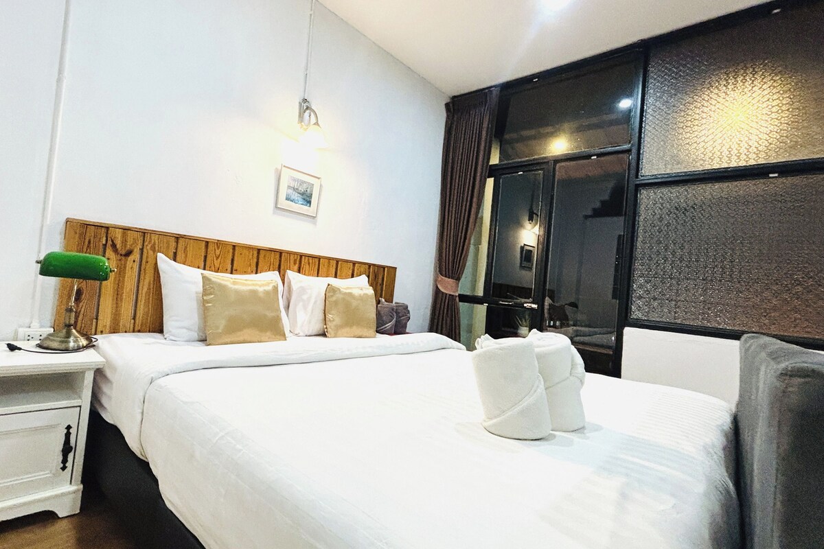 Uma cama bem arrumada com lençóis brancos, cabeceira de madeira, abajur e uma janela com cortinas.