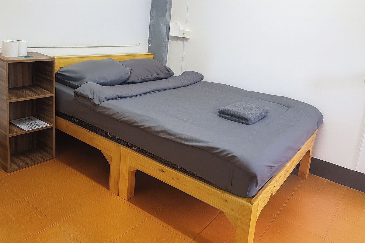 Un lit simple avec un cadre en bois, une literie grise et une petite étagère à côté.