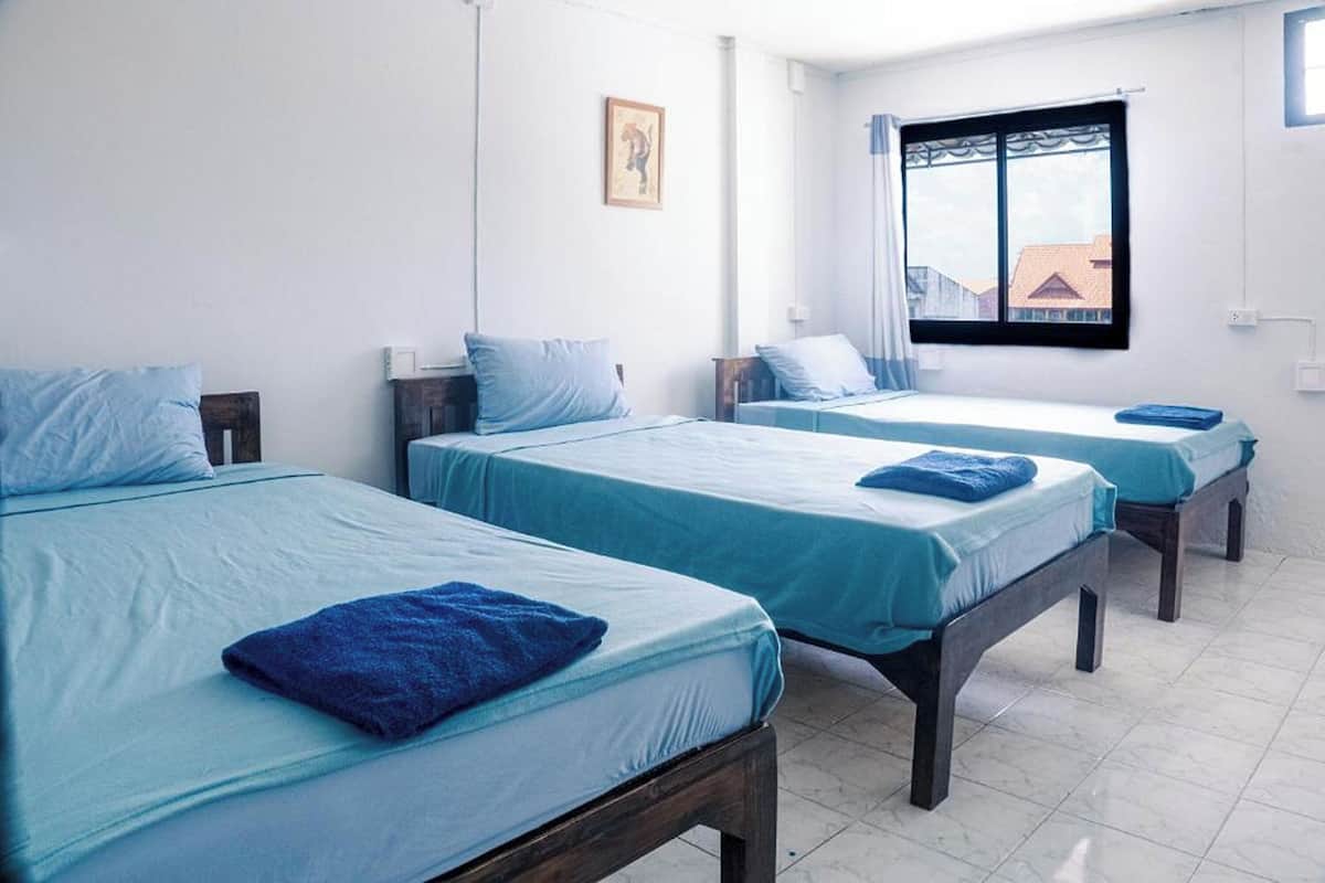 Une chambre d’hôtel avec trois lits simples, chacun recouvert d’une couverture bleue et doté d’un oreiller, une fenêtre donnant sur des immeubles et un mur blanc sobre orné d’un tableau encadré.