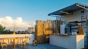 Un espace extérieur aménagé avec une table et des tabourets en bois, une clôture en bambou et un petit comptoir de bar agrémenté d’une enseigne indiquant le prix d’un verre et d’une bière.