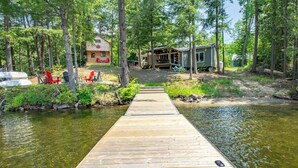 A wooden dock leading to a cabin with a red roof and a maple leaf emblem, surrounded by trees and a small docked boat. - Steenburg Shores & S'mores (Gilmour)