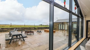 A patio area with picnic tables and a stone floor, overlooking a grassy field and a building with a sloped roof. - Highgate House (Dronfield)