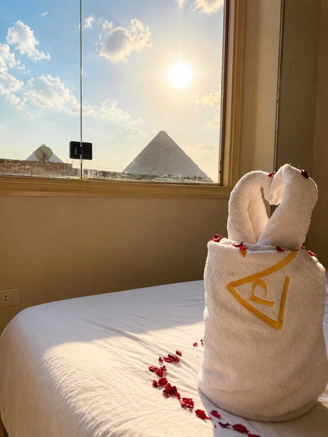 A hotel room with a bed, a window with a view of the pyramids, and a towel with a symbol on it.