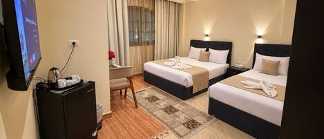 A hotel room with two beds, a desk with a computer and coffee maker, a TV, and a large window with curtains.
