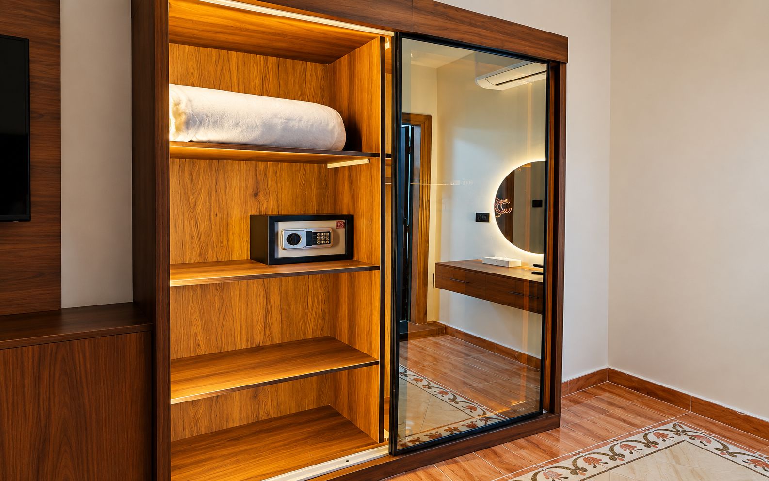 A wooden wardrobe with a mirror, a safe, and a shelf with a rolled-up towel.
