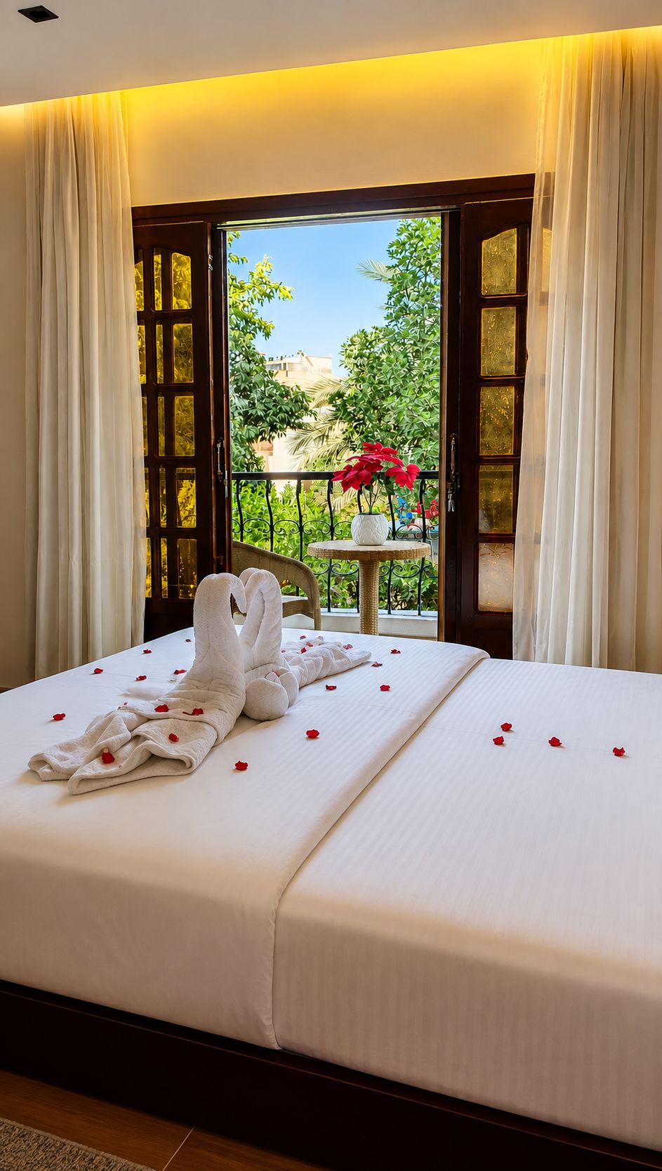 A hotel room with a bed, a view of greenery outside, and a table with a vase of flowers.