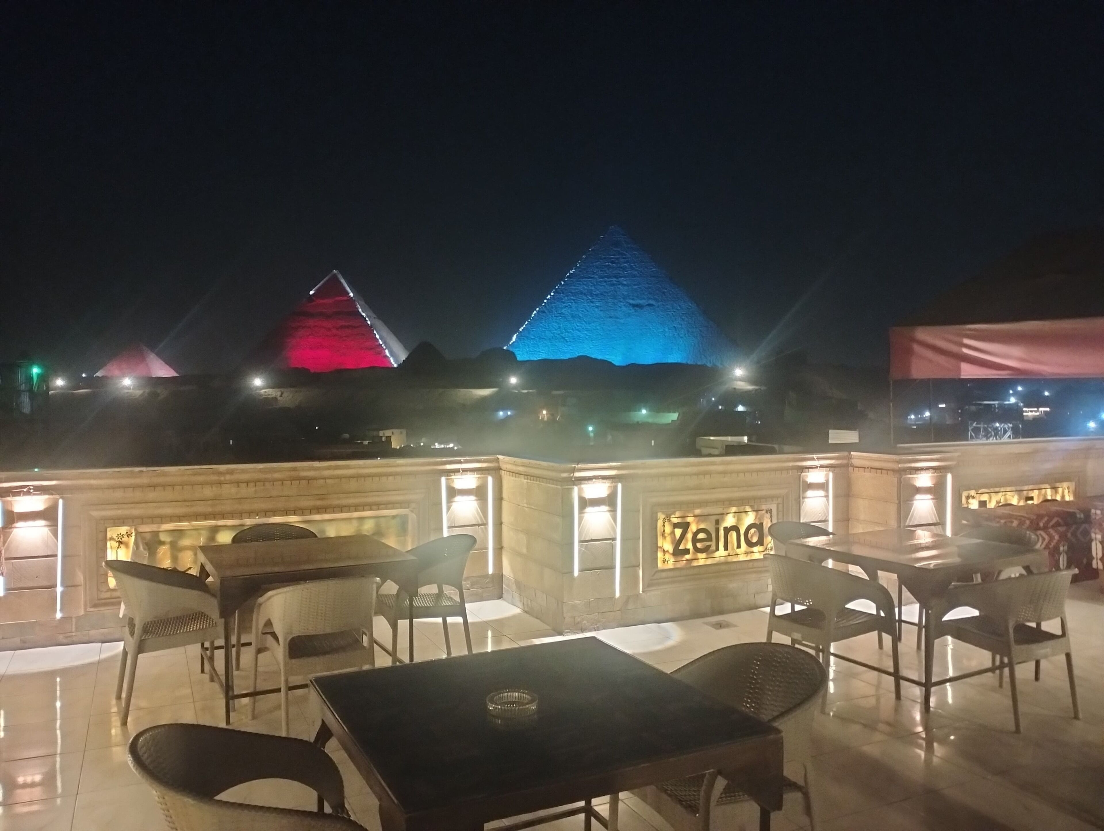 An outdoor dining area with tables and chairs, a sign reading "Zeina," and illuminated pyramids in the background.