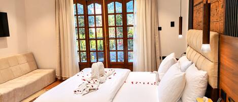 A hotel room with a bed, a sofa, a wooden headboard, and a view through a large window.