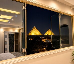 A room with a large window overlooking two illuminated pyramids at night. - Pyramids Kingdom RZ Hotel (Giza)