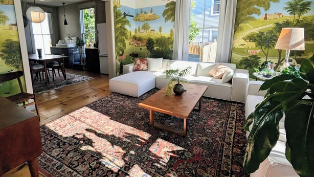 A living room with a sofa, coffee table, and a large rug. There is a dining area with chairs and a table. The room has a mural on the wall depicting a landscape. - Chic 3-bedroom house in Munjoy Hill with off-street parking.   (Portland)