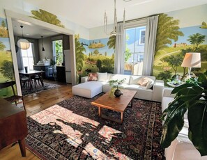 A living room with a sofa, coffee table, and a large rug. There is a dining area with chairs and a table. The room has a mural on the wall depicting a landscape. - Chic 3-bedroom house in Munjoy Hill with off-street parking.   (Portland)