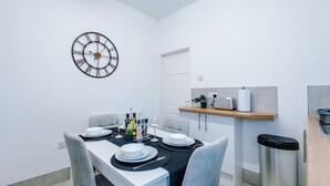 Un coin repas avec une horloge murale ronde, une table blanche dressée pour quatre personnes, des chaises grises, une petite plante et une poubelle.