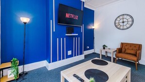Une pièce avec un mur d’accent bleu, un téléviseur fixé au mur, une horloge, un fauteuil marron, une table basse et un lampadaire.