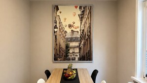 Uma área de refeições com mesa de madeira, cadeiras, um jogo americano preto e uma pintura de paisagem urbana na parede.