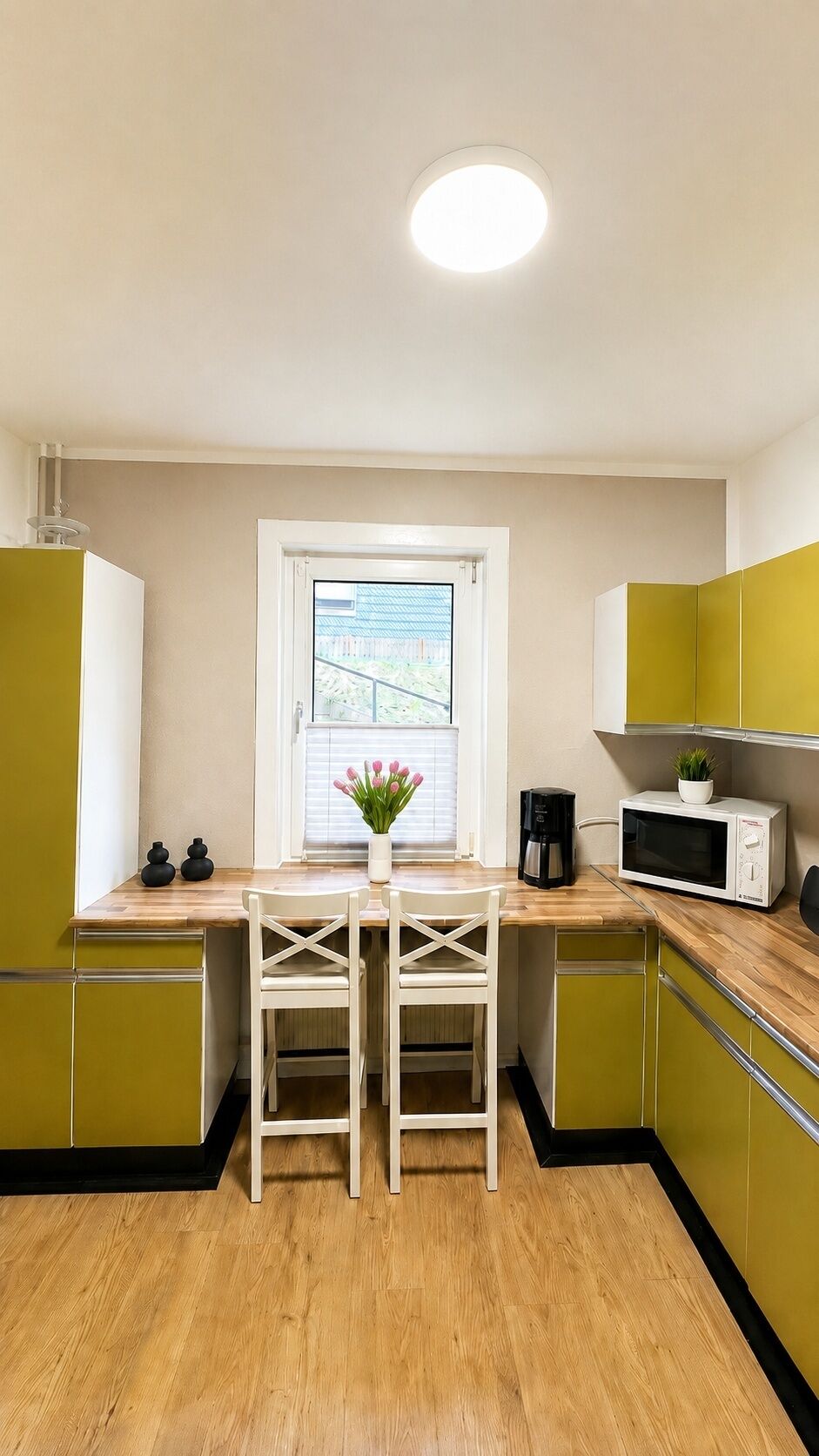 A compact kitchen with a wooden countertop, a white chair, a window with a view, and green cabinets.