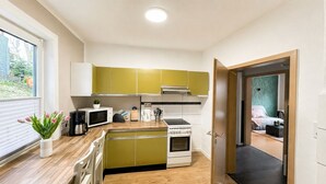 Uma cozinha compacta com micro-ondas, fogão e pia, uma pequena área de refeições com uma cadeira e vista para um quarto por uma porta aberta.