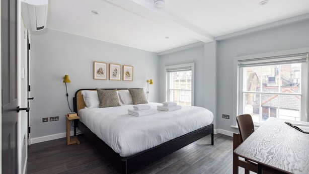 A modern bedroom with a large bed, bedside tables, a dining table, and a window with a city view. - Hausd - Soho (London)