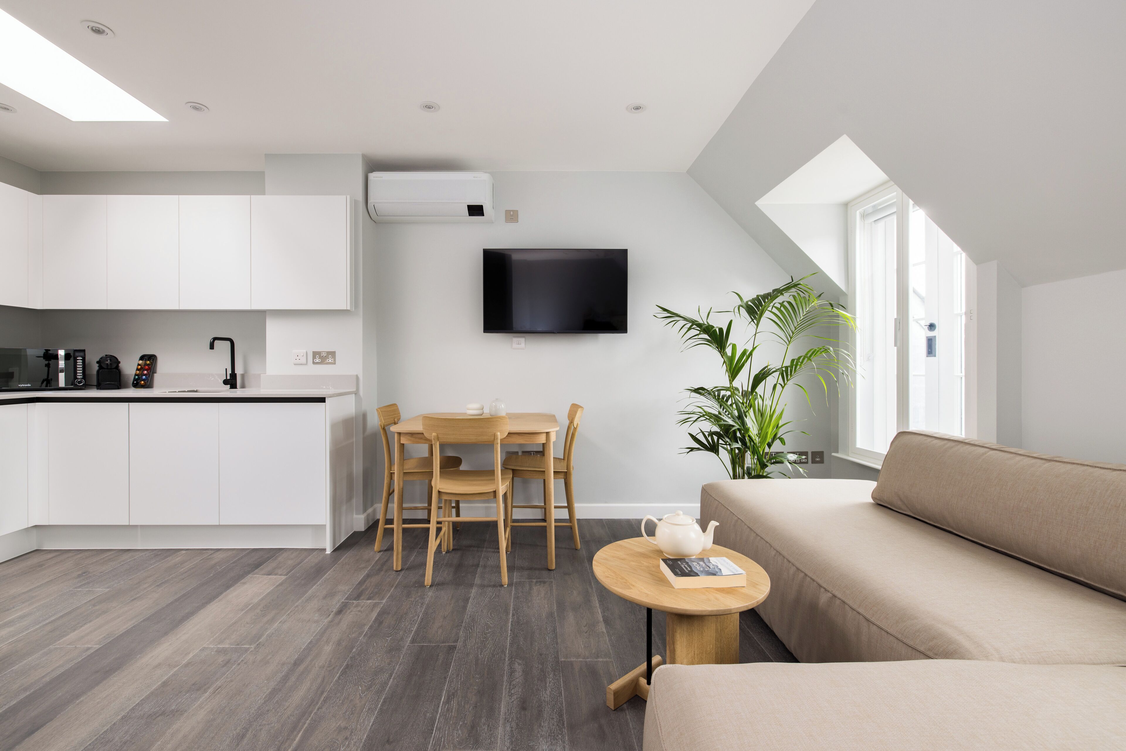 A modern kitchen with white cabinets, a dining area with a wooden table and chairs, a beige sofa, and a flat-screen TV mounted on the wall.
