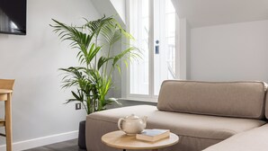 A modern living room with a beige sofa, a round wooden table with a teapot and book, and a large green plant. - Hausd - Soho (London)