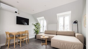 A modern living room with a sectional sofa, a dining table with chairs, a small side table, a potted plant, and a flat-screen TV mounted on the wall. - Hausd - Soho (London)