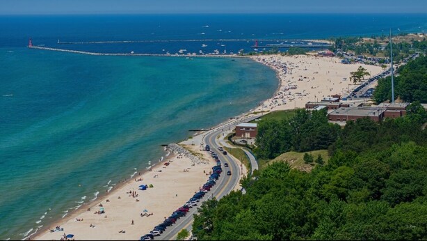 Une vue aérienne d’une plage, avec une longue étendue de sable, une route parallèle au rivage et un phare visible au loin.