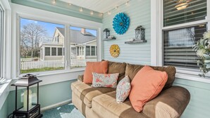 Une pièce chaleureuse avec un canapé beige, des coussins décoratifs et une vue sur une maison, visible à travers une grande fenêtre.