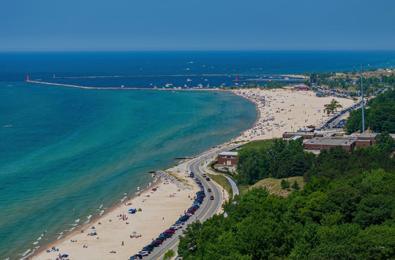 An aerial view of a beach with a long stretch of beach, a road parallel to the shore, and a lighthouse in the distance.