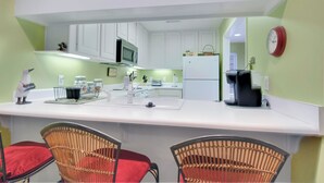 A kitchen with white cabinets, a microwave, a refrigerator, and a coffee maker. There are wicker chairs with red cushions and a white countertop.
