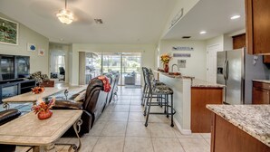 A spacious living room with a large TV, a dining area with a table and chairs, and a kitchen with a refrigerator and cabinets. - 4 Bedroom Fresh Water Canal Home with Saltwater Pool and Hot Tub! (Cape Coral, FL)