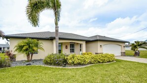 A single-story house with a beige exterior, a grey roof, and a well-maintained front yard with grass and various plants. - 4 Bedroom Fresh Water Canal Home with Saltwater Pool and Hot Tub! (Cape Coral, FL)