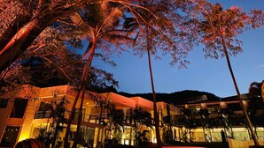 Un edificio de varios pisos con balcón y zona de estar al aire libre, rodeado de palmeras bajo un cielo nocturno.