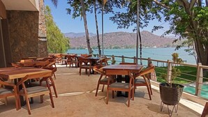 Un espacio al aire libre con mesas y sillas de madera, un muro de piedra y vistas a un lago con montañas al fondo.